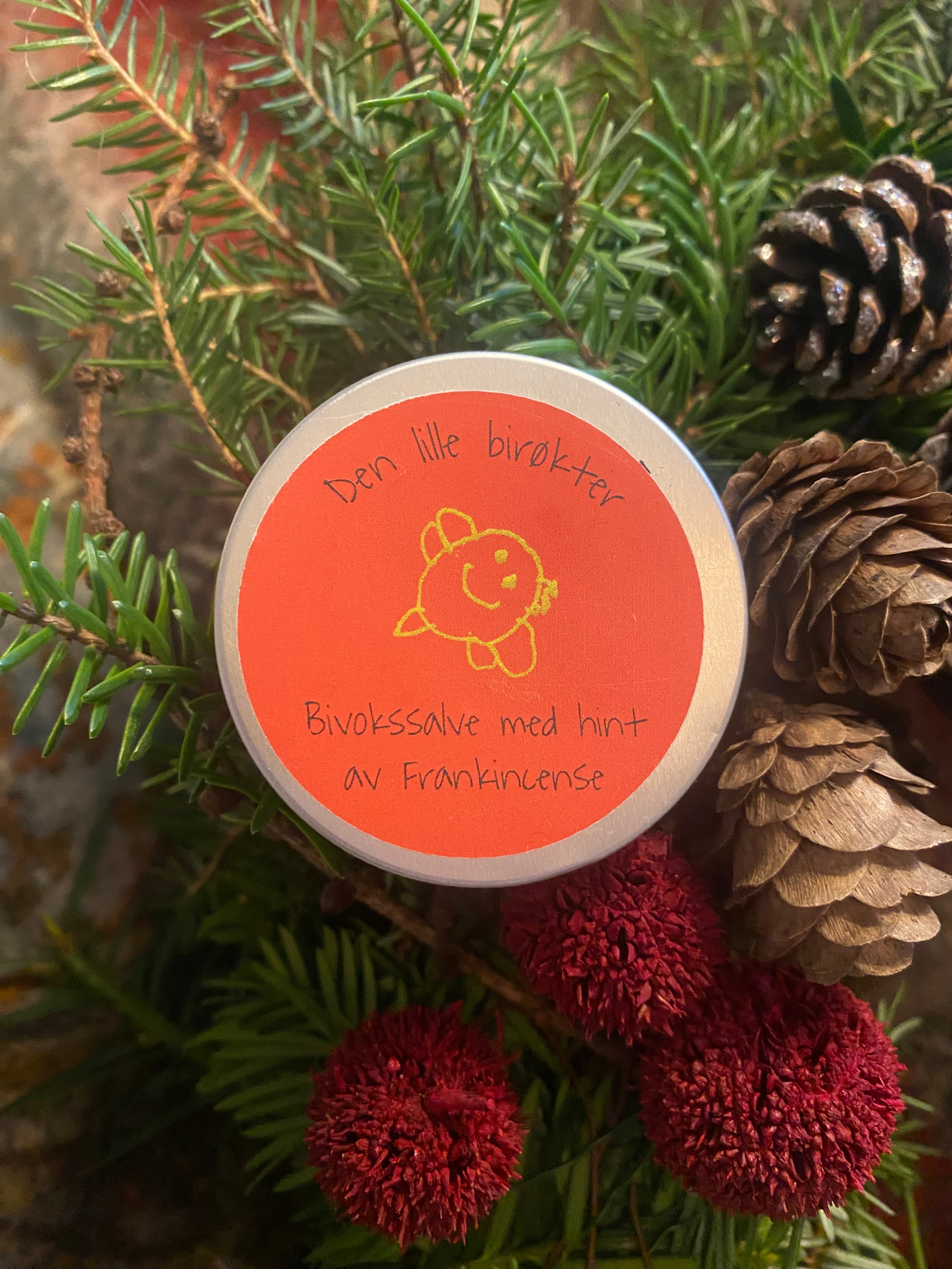 Liten aluminiumsboks med rød bakgrunn og gullbie som ligger på en krans med vintergrønne kvister og kongler