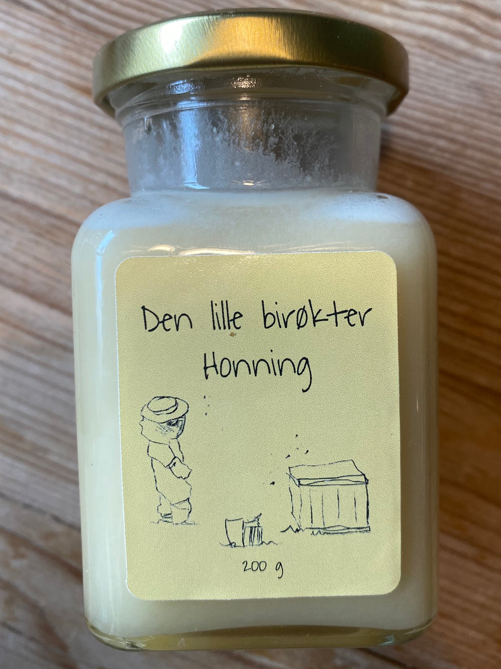 Honningglass med lys gul etikett på furubakgrunn. Bilde av liten birøkter ved siden av en bikube.