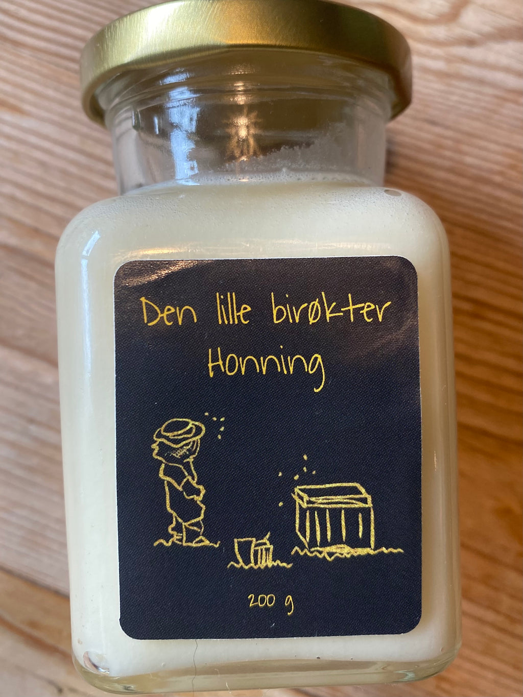 Honningglass med svart etikett liggende på furubakgrunn. Bilde av liten birøkter og bikube tegnet i gull.